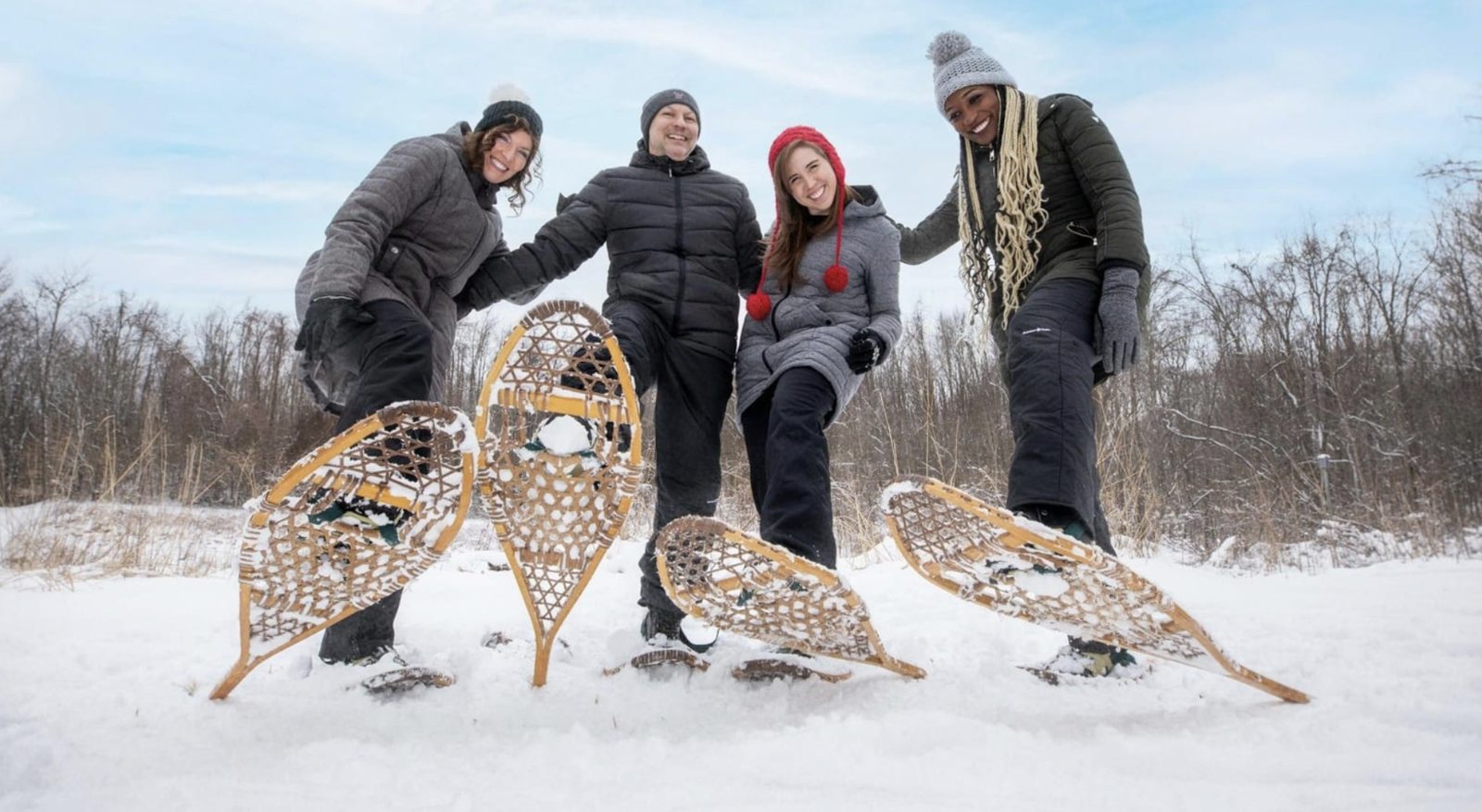 Smiling group on snowshoes in fresh snow, lifting their snowshoes towards the camera