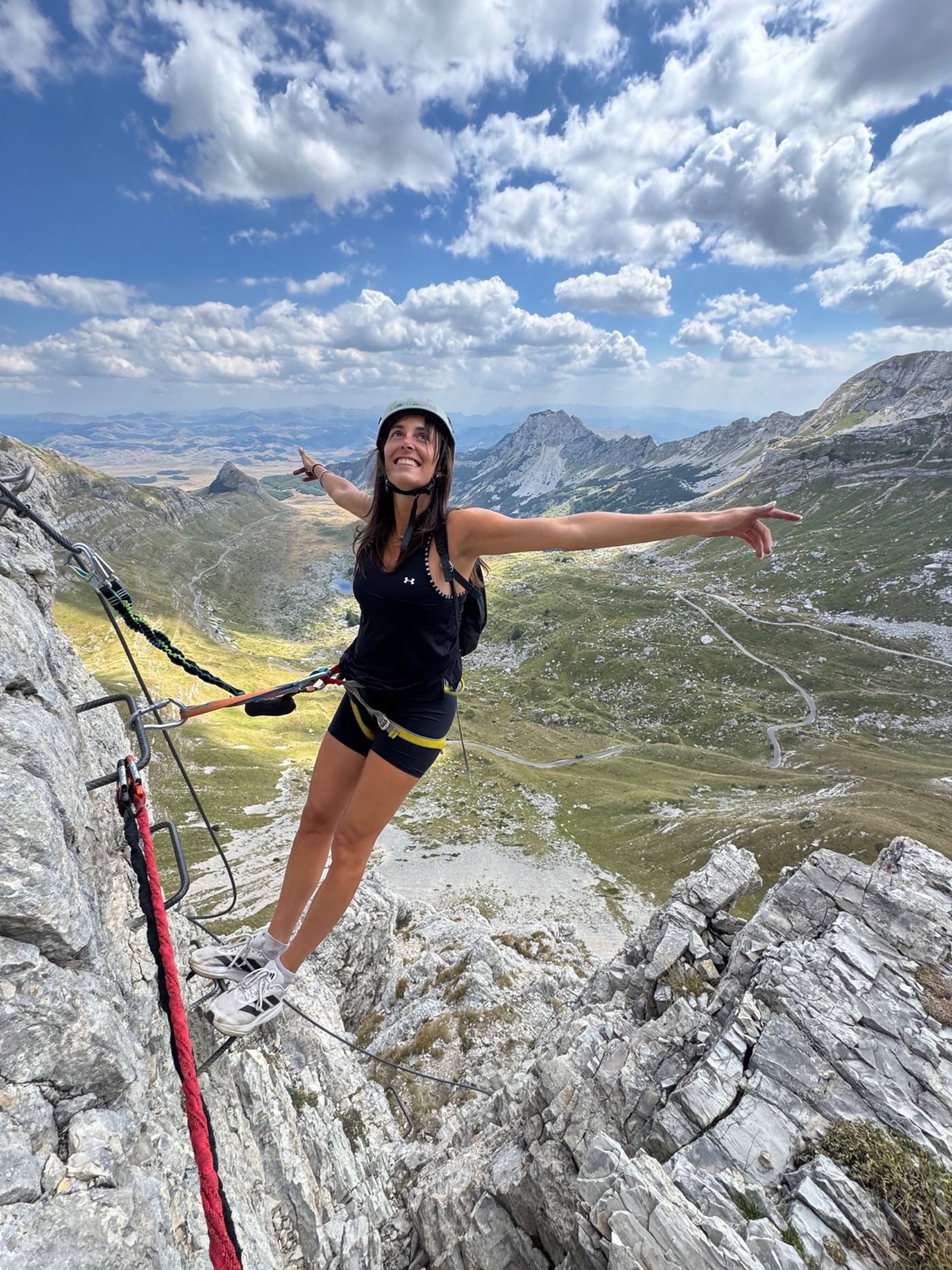 Smiling climber clipped to the steel cable on the Durmitor via ferrata, arms spread above the green valley and Sedlo road